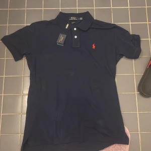 Marinblå pikétröja Polo Ralph Lauren - Snygg marinblå pikétröja från Polo Ralph Lauren i custom fit. Klassisk design med krage, tre knappar och röd broderad logga på bröstet. Tillverkad i mjuk bomull som känns skön mot huden. Perfekt för en clean och stilren look.
