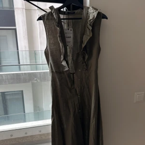 Olivgrön ärmlös klänning från Zara midi  - Snygg olivgrön klänning från Zara med volangdetaljer längs v-ringningen och knappar framtill. Ärmlös modell i glansigt material som ger en cool look. Perfekt för dig som gillar stilrena men unika plagg. 