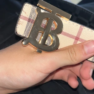 Burberry bälte med rutigt mönster - Säljer ett Burberry bälte med klassiskt rutigt mönster i beige, svart och rött på ena sidan och svart rutigt på andra. Snyggt guldfärgat spänne med Burberrys ikoniska TB-logo. Bältet är gjort i skinn och har en exklusiv känsla.