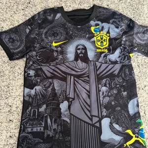 Brasil Nike fotbollströja svart/grå - Unik Brasilien fotbollströja från Nike med svart och grått konstnärligt tryck av Kristusstatyn och brasilianska motiv. Gula detaljer, CBF-emblem på bröstet och korta ärmar. Tillverkad i lätt och ventilerande material, perfekt för match eller träning.