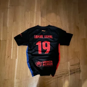 FC Barcelona Lamine Yamal tröja svart - Snygg svart FC Barcelona fotbollströja med Lamine Yamal och nummer 19 på ryggen. Tröjan har röda detaljer, UNHCR-logga och klubbmärke på bröstet. Sidorna har blå och röda inslag. Perfekt för dig som vill visa ditt stöd för Barça och Yamal.