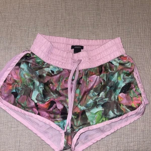 Träningsshorts från lindex - Snygga träningsshorts från Lindex i storlek XS. De har ett blommigt mönster i lila, grönt och rosa med ljuslila resårmidja och snörning. Perfekta för sommarens aktiviteter och riktigt bekväma med loose fit och mesh-detaljer på sidorna.