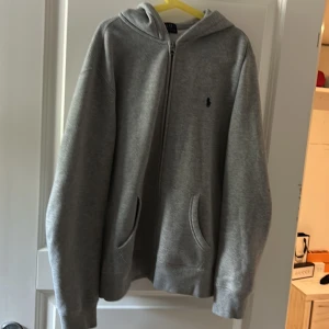 Grå hoodie från Polo Ralph Lauren.storlek är 14 till 16 år  - Äkta Klassisk grå hoodie från Polo Ralph Lauren  med dragkedja framtill och broderad logga på bröstet. Tröjan har huva, två fickor och ribbade muddar vid ärmslut och nederkant. Perfekt för chill dagar och enkel att matcha med allt.