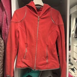 Röd hoodiejacka från Belstaff - Röd hoodiejacka från Belstaff med stora broderade loggor på ryggen och coola kontrastsömmar. Jackan har dragkedja framtill, två fickor med dragkedja och en sportig vibe. 