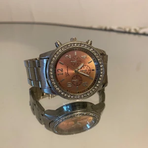 Silverfärgad klocka med strass från Geneva - Stilren klocka från Geneva med silverfärgat armband i metall och en skimrande roséguldfärgad urtavla. Runt urtavlan sitter glittrande strass-stenar som ger extra bling. Klockan har tre dekorativa små urtavlor och tydliga siffror.