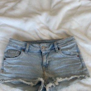 Ljusblå jeansshorts med fransig kant - Snygga ljusblå jeansshorts med fransig, rå kant nertill och klassisk femficksmodell. Shortsen har låg midja och dragkedja med knapp framtill. Perfekta för varma sommardagar.