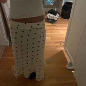 Vit prickig långkjol med volang - Superfin vit långkjol med bruna prickar och volang nertill. Kjolen har en rak passform som sitter snyggt i midjan och ger en chill men ändå söt vibe. Perfekt för dig som gillar lekfulla mönster och vill sticka ut lite extra.