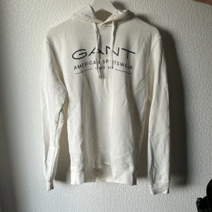 Vit hoodie från GANT med tryck - Vit hoodie från GANT med svart tryck på bröstet där det står 'GANT American Sportswear'. Tröjan har huva med snörning och långa ärmar. Mjuk och skön bomullskänsla, perfekt för en chill och stilren look.