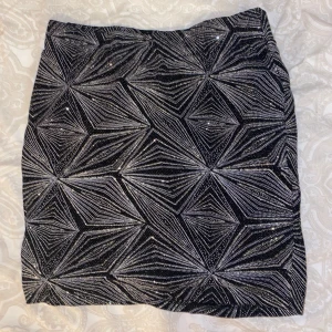 Glittrig svart och silver kortkjol - Snygg kortkjol i svart med glittrigt silvermönster i geometrisk stil. Kjolen har en tajt passform och är perfekt för dig som vill sticka ut. Materialet känns festligt och skimrande.