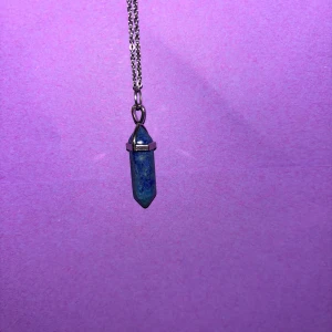 Halsband med blå Lapis Lazuli-sten - Snyggt halsband med en spetsformad blå Lapis Lazuli-sten som hänge. Kedjan är i silverfärg och stenen har naturliga mönster i olika blå nyanser. Perfekt för dig som gillar kristaller och unika smycken.
