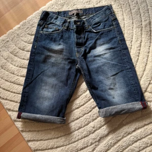 Blå jeansshorts från L.O.G.G. - Snygga blå jeansshorts från L.O.G.G. med klassisk femficksdesign och uppvikta benslut. Shortsen har en mörk tvätt med lätt slitning och kontrastsömmar. Perfekta för en chill sommarstil och funkar till allt från t-shirt till hoodie.
