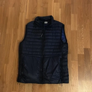 Mörkblå väst från C.P. Company - Snygg mörkblå dunväst från C.P. Company med quiltad design och hög krage. Västen har dragkedja framtill och två fickor med dragkedja. Tillverkad i syntetmaterial med dunfyllning, perfekt för lager-på-lager-stil. D.D. Shell-modell. Köpt på NK, nypris 4200