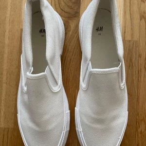 Vita slip-on sneakers - Säljer ett par klassiska vita slip-on sneakers från H&M i storlek 40. Skorna har en platt sula och är tillverkade i canvasmaterial. De har vaderad kant, resår i sidorna och hälla bak. Foder och innersula i bomullscanvas. Använda en gång och är i mycket fint skick.