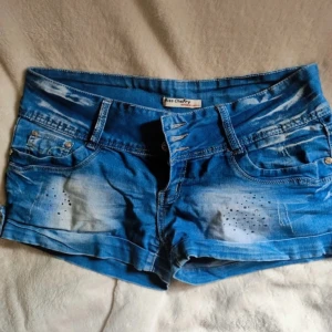 Blå jeansshorts från Miss Cherry - Snygga blå jeansshorts från Miss Cherry med dekorativa slitningar och små nitar framtill. Shortsen har normal passform, klassiska fickor fram och bak samt knäppning med tre knappar 