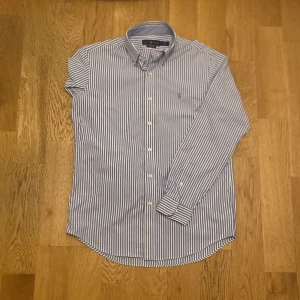 Blåvit randig skjorta från Ralph Lauren - Klassisk blåvit randig skjorta från Ralph Lauren med broderad logga på bröstet. Skjortan har lång ärm, knappar framtill och är tillverkad i ett mjukt bomullstyg. Perfekt för en stilren och tidlös look.