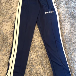 Blå träningsbyxor från Palm Angels - Snygga blå träningsbyxor från Palm Angels med vita sidoränder och logga tryckt på ena benet. Byxorna har elastisk midja med dragsko och raka ben. Perfekta för en sportig och trendig look.