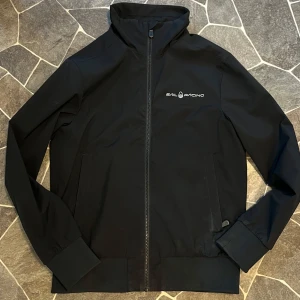 Svart jacka från Sail Racing - Säljer en svart jacka från Sail Racing med hög krage och dragkedja framtill. Jackan har två sidofickor och en diskret logga på bröstet samt en detalj på ryggen. Perfekt för dig som gillar stilren design.