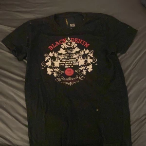 Svart t-shirt med tryck från Jack & Jones - Svart t-shirt från Jack & Jones med stort tryck på bröstet i rött och vitt. Texten 'Black Denim' och dekorativa detaljer ger en cool och stilren look. Perfekt till jeans eller shorts. Smaller sized M