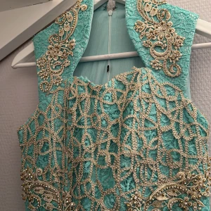 Turkos långklänning med spets och guldbroderi - Säljer en turkos långklänning med vackra guldfärgade broderier och spetsdetaljer. Klänningen har hög krage och är ärmlös, med dragkedja i ryggen. Perfekt för speciella tillfällen när du vill sticka ut.