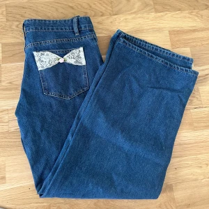 Blå jeans med spetsdetaljer från Edikted - Säljer ett par blå jeans från Edikted med unika spetsdetaljer och små rosetter på bakfickorna. Klassisk femficksmodell med dragkedja och knapp framtill. Perfekta för dig som vill ha något extra på dina jeans! Helt nya med prisplappen på, säljes pga fel storlek. 