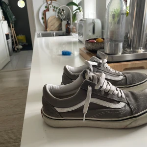 Gråa Vans Old Skool sneakers - Säljer ett par klassiska Vans Old Skool sneakers i grå mocka med vita detaljer och vit sula. Skorna har den ikoniska vita sidoremsan och snörning framtill. Perfekta för en avslappnad och stilren look.