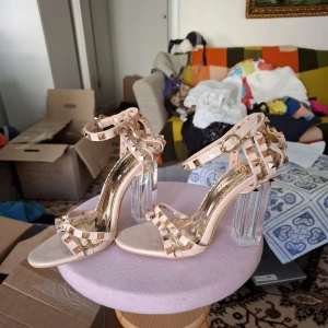 Beige klackskor med nitar och transparent klack - Säljer ett par snygga beige klackskor med remmar dekorerade med guldiga nitar. Skorna har en hög, transparent blockklack och öppen tå. Perfekta för dig som vill sticka ut med en trendig och edgy look.