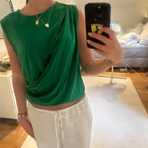 Grön draperad ärmlös topp - Säljer en snygg grön ärmlös topp med draperad detalj framtill. Toppen har en rund halsringning och är tillverkad i ett mjukt, lätt material som ger en avslappnad men trendig look.