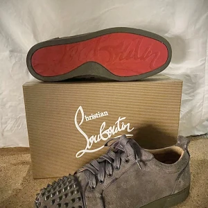 Gråa sneakers från Christian Louboutin med nitar - Säljer ett par gråa sneakers från Christian Louboutin med klassisk röd sula och coola nitar framtill. Skorna har snörning och är tillverkade i mocka för en lyxig känsla. Perfekta för dig som vill sticka ut.