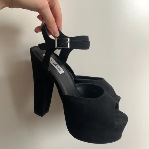 Svarta platåklackar med öppen tå från NLY shoes - Säljer ett par svarta platåklackar med öppen tå och hög klack. Skorna har en justerbar rem med spänne runt ankeln och är tillverkade i mocka. Perfekta för dig som vill ha extra höjd och stabilitet.