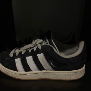 Äkta Adidas Campus sneakers  - Säljer ett par klassiska Adidas Campus sneakers i svart mocka med vita ränder och vit sula. Skorna har snörning och den ikoniska Adidas-loggan på hälen. Perfekta för en stilren och sportig look.