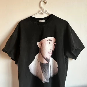 Svart t-shirt med tryck från Ask Yourself - Cool svart t-shirt med stort porträtttryck framtill och en stor vit korsdetalj med texten 'Ask Yourself' på ryggen. Klassisk passform och rund halsringning. Perfekt för dig som gillar streetwear och statement-plagg.