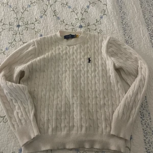 Vit kabelstickad tröja från Polo Ralph Lauren - Kabelstickad vit tröja från Polo Ralph Lauren med rund halsringning och broderad logga på bröstet. Klassisk modell med ribbade muddar vid ärmslut och nederkant. Perfekt för en stilren och tidlös look. Den är liten i storlek passar s och xs