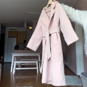 Ljusrosa kappa med bälte - Säljer en ljusrosa kappa med klassisk krage och bälte i midjan. Kappan har långa ärmar, knappar framtill och två fickor. Perfekt för dig som vill ha en stilren och feminin look. 100%wool 