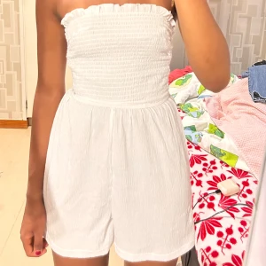 Vit axelbandslös playsuit - Supersöt vit axelbandslös playsuit med smock upptill och insydda shorts. Lätt och luftig modell med korta ben, 