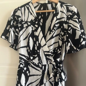 Svartvit omlottklänning med knyt - Säljer en svartvit omlottklänning med grafiskt mönster och knyt i midjan. Klänningen har korta ärmar och v-ringad hals. Perfekt för dig som gillar en stilren men ändå iögonfallande look. Lite över knäna