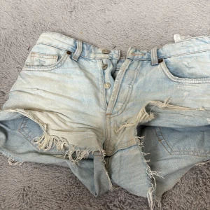 Ljusblå jeansshorts med fransar från ZARA - Säljer ett par ljusblå jeansshorts från ZARA med fransiga kanter och slitna detaljer. Shortsen har klassisk femficksmodell, knappgylf och bälteshällor. Perfekta för en avslappnad stil.