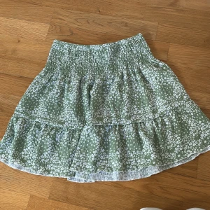 Grön blommig kjol från Gina Tricot - Supersöt grön kjol från Gina Tricot med vitt blommönster och volang. Kjolen har smock i midjan och är kort i modellen, perfekt för varma dagar. Lätt och luftig känsla med härligt fall. Kan man också använda som tröja.
