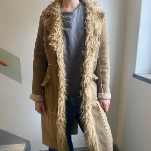 Beige lång pälsjacka från Vero Moda - Lång beige pälsjacka från Vero Moda med fluffig fuskpäls längs krage och öppning. Jackan har markerade sömmar, bred krage och uppvikta ärmslut. En klassiker!