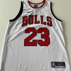 Vit Chicago Bulls basketlinne  - Säljer ett vitt Chicago Bulls basketlinne, nummer 23 och Jordan på ryggen. Klassisk NBA-design med svarta detaljer vid ärmhålerna och halsen.