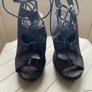 Svarta klackskor med snörning från Graceland - Säljer ett par svarta klackskor från Graceland med öppen tå och snörning framtill. Skorna har hög klack, grov sula och är tillverkade i ett material som liknar mocka. Perfekta för dig som vill sticka ut med en edgy stil.