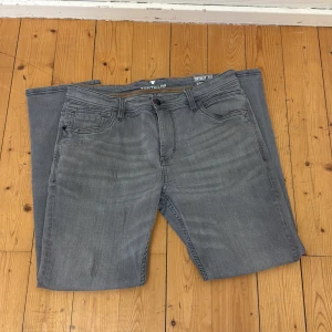 Grå jeans från Tom Tailor Josh Regular Slim - Säljer ett par grå jeans från Tom Tailor, modell Josh Regular Slim. Byxorna har klassisk femficksdesign, orangea sömmar och en slim passform. Perfekta för dig som gillar en stilren och enkel look. 