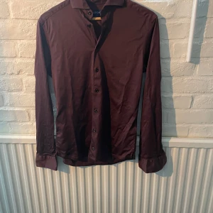 Eton skjorta M - Säljer en stilren mörkröd skjorta från Eton i slim fit. Skjortan har långa ärmar, klassisk krage och knäppning framtill. Perfekt för dig som vill ha en snygg och tidlös look. Storlek M 