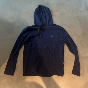 Mörkblå hoodie från Ralph Lauren - Mörkblå hoodie från Ralph Lauren med liten broderad logga på bröstet. Tröjan har långa ärmar och huva med dragsko. Perfekt för en avslappnad stil. Ganska tunn perfekt till sommaren