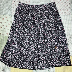 Svart blommig kjol från Ü - Säljer en svart kjol från Ü med blommigt mönster i vitt och rosa. Kjolen har resår i midjan och volang nertill för en söt och avslappnad look. Perfekt för dig som gillar mjuka material och fina detaljer. Nästan Aldrig andvänd.