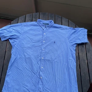 Blårandig skjorta från Ralph Lauren - Klassisk blårandig skjorta från Ralph Lauren med broderad logga på bröstet. Skjortan har korta ärmar, knappar framtill och är tillverkad i 100% bomull. Perfekt för en stilren och avslappnad look. Nytvättad, vi stryker den innan vi fraktar den till dig!