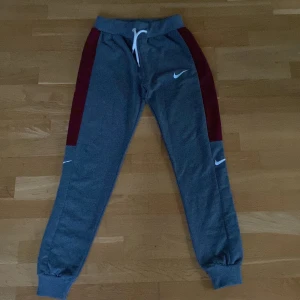 Grå mjukisbyxor från Nike med röda detaljer - Snygga grå mjukisbyxor från Nike med röda paneler längs sidorna och vita Nike-loggor. Byxorna har resår och snörning i midjan samt muddar vid bensluten. Perfekta för en avslappnad stil. Storlek är M för barn.