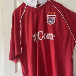 Bayern München fotbollströja Makaay 10 retro - Röd Bayern München fotbollströja med vita detaljer, T-Com sponsortryck och klubbmärke på bröstet. Nummer 10 och Makaay på ryggen. Klassisk krage och korta ärmar. Perfekt för fans av tysk fotboll.