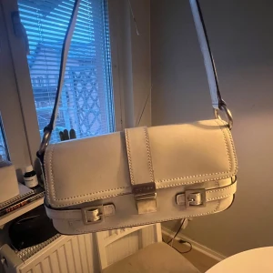 Vit axelväska från Guess - Säljer en stilren vit axelväska från Guess med silverfärgade metalldetaljer och spänne framtill. Väskan har en rektangulär form och justerbar axelrem. Perfekt för att lyfta din outfit med en klassisk touch.