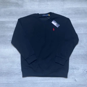 Svart sweatshirt från Polo Ralph Lauren - Klassisk svart sweatshirt från Polo Ralph Lauren med rund halsringning och röd broderad logga på bröstet. Tröjan har långa ärmar och ribbade muddar vid ärmslut och nederkant. Perfekt för en stilren och enkel look.