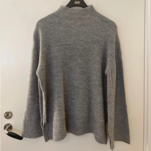 Grå stickad polotröja från 157 - Mysig grå stickad polotröja från 157 med ribbad struktur. Tröjan har lång ärm, hög krage och slitsar i sidorna för en avslappnad look. Perfekt för lager-på-lager under kyliga dagar.